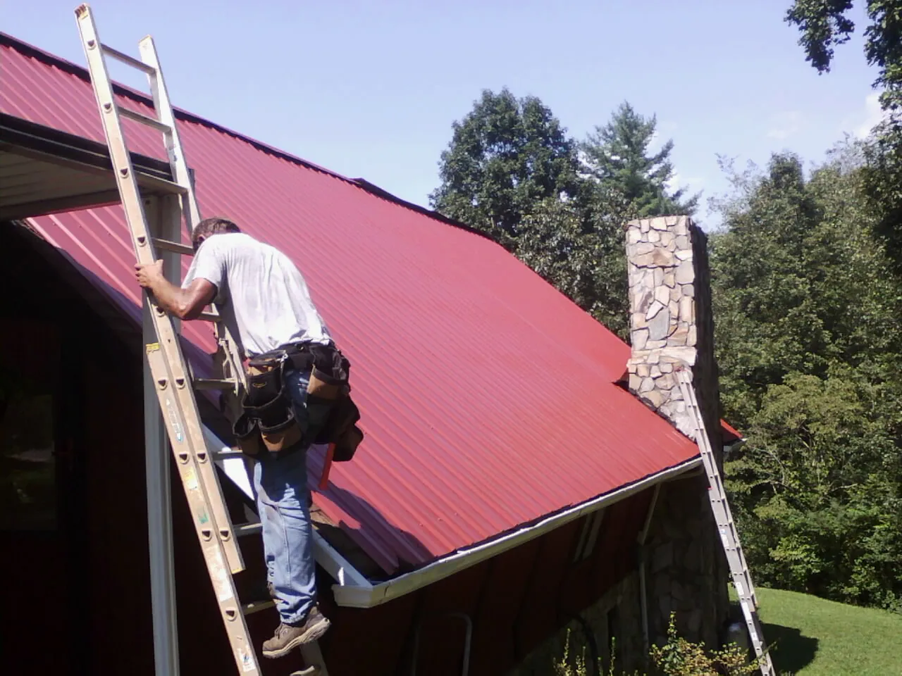Roofer on a ladder beside a red metal roof and stone chimney — Soffit & Fascia Repair by Roofing Brothers in Mc Donald